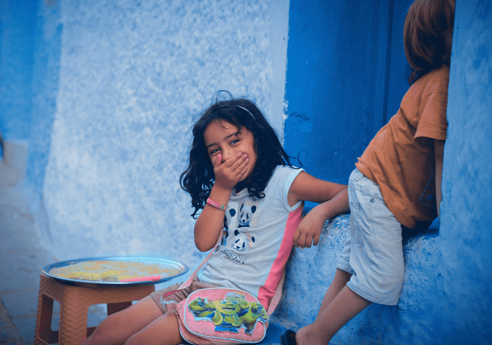 Girl giggling behind her hand outside blue house in Morocco