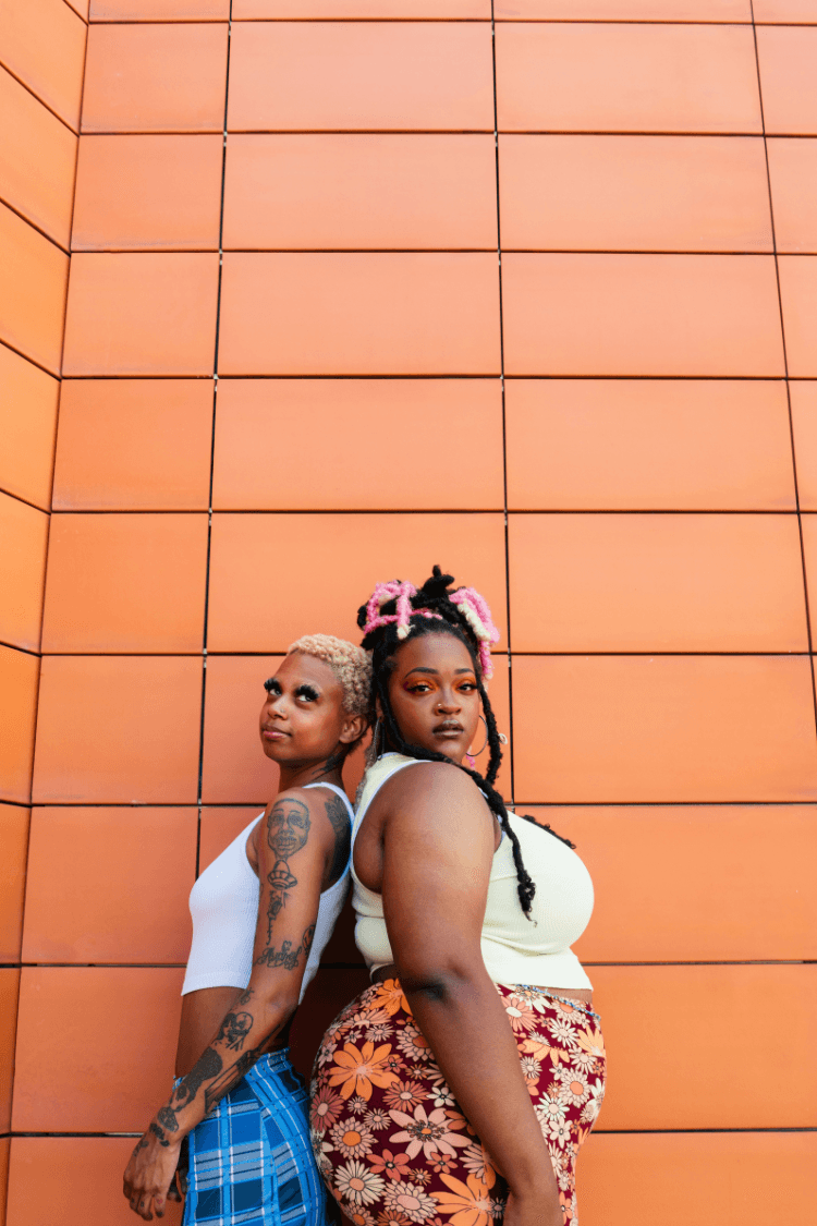 Two women posing back to back in front of an orange wall
