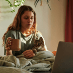 Woman sitting in bed looking at laptop holding mug and cat