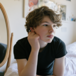 A teenage boy sitting by a bedroom mirror looking pensive
